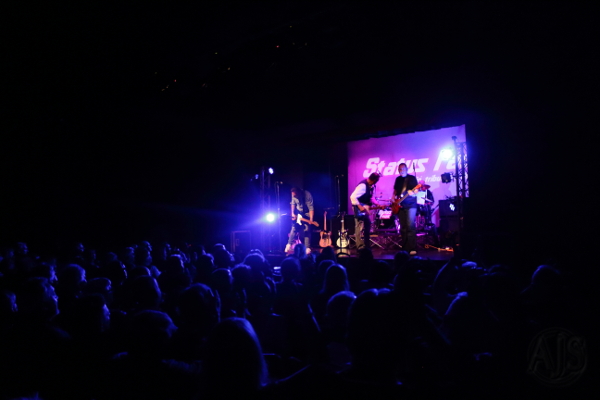 The band in a darkened auditorium with a single light shining out across the audience.