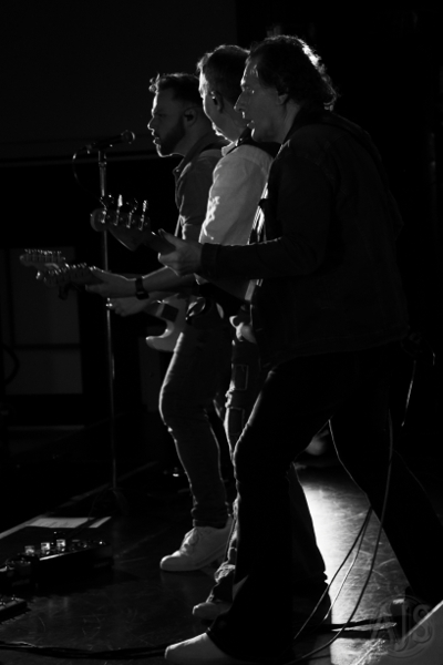 The three Status Faux guitarists on stage together in profile. Black and white.