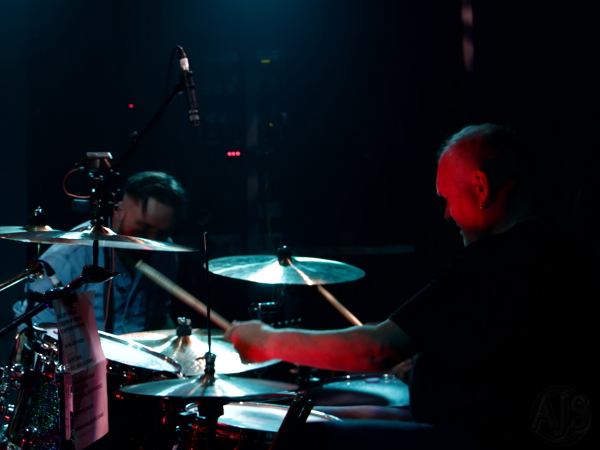 Drummer playing on stage with drum kit softly lit and guitarist leaning towards him.