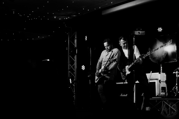 The Status Faux frontmen performing together on a dark stage highlighted by a single light.